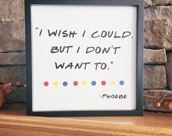 Op de afbeelding: Een zwart ingelijst print met een witte achtergrond en de quote "Ik wou dat ik kon, maar ik wil niet." in zwarte handgeschreven letter. De quote wordt toegeschreven aan "Phoebe" in een kleiner lettertype. Er zijn negen kleurrijke stippen in een rij onder de quote.