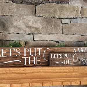 May include: Two wooden signs with the text "Let's Put The Crazy Away" stacked on top of each other. The signs are brown with white lettering and are sitting on a wooden mantel.