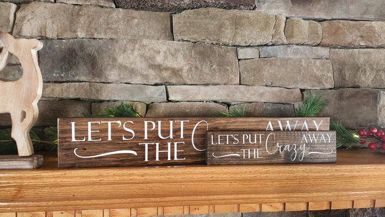 May include: Two wooden signs with the text "Let's Put The Crazy Away" stacked on top of each other. The signs are brown with white lettering and are sitting on a wooden mantel.