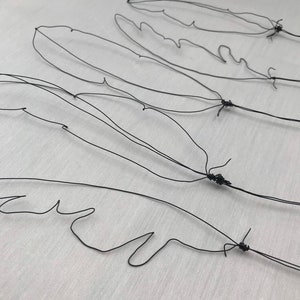May include: Four black wire feather sculptures are arranged on a white surface. Each feather is crafted from a single wire, with detailed vein-like structures. The feathers are tied together at the base.