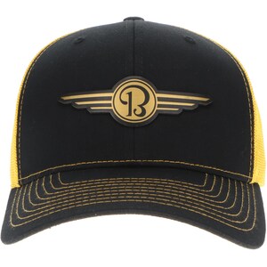 Könnte beinhalten: Eine schwarze Baseballkappe mit einem goldfarbenen Emblem auf der Vorderseite, das einen stilisierten Buchstaben B in einem geflügelten Design zeigt. Die Kappe hat eine strukturierte Krone, einen gebogenen Schirm mit goldfarbenen Nähten sowie gelbe Netz-Seitenteile.