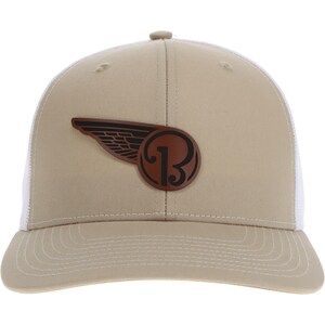 Könnte beinhalten: Eine beige Baseballkappe mit weißem Netzrücken und gebogenem Schirm. Auf der Vorderseite befindet sich ein kreisförmiger brauner Aufnäher mit einem schwarzen, stilisierten Buchstaben B und einem Flügeldesign. Die Kappe hat einen Knopf und Nähte.
