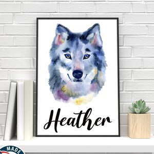 May include: A framed watercolor print of a wolf's face, predominantly blue and gray, with yellow eyes. The name "Heather" is written in black script below the image. The print is displayed on a white shelf with books and a small succulent.
