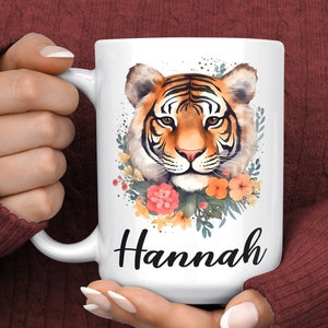 May include: White ceramic mug with a watercolor illustration of a tiger's face surrounded by flowers. The name "Hannah" is printed in black below the image.
