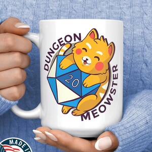 May include: White ceramic mug with a cartoon cat holding a blue 20-sided die. The text "Dungeon Meowster" is printed around the die.