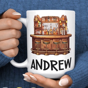 May include: White ceramic mug with a bar scene illustration and the name "ANDREW" printed in black below. The bar scene features a wooden counter with stools and shelves stocked with bottles of liquor.