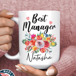May include: White ceramic mug with a floral design and the text "Best Manager Natasha" in black lettering. The mug has pink hearts on the top and bottom of the text.