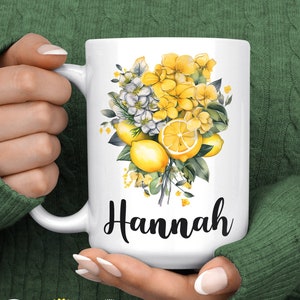 May include: White ceramic mug featuring a floral design with lemons and yellow flowers. The name "Hannah" is written in black script. The mug is held by a person wearing a green sweater.
