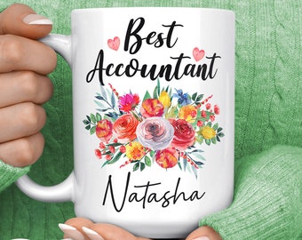 Op de afbeelding: Witte keramische mok met de tekst "Best Accountant Natasha" in zwart schrift, versierd met een bloemenboeket van rode, oranje, gele en blauwe bloemen. De mok wordt vastgehouden door een persoon die een groene trui draagt. Een "Made in USA" sticker is zichtbaar.