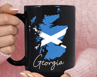 May include: Black coffee mug with a white outline of the state of Georgia and the word "Georgia" written in cursive. The state of Georgia is filled in with the colors of the Scottish flag, a blue field with a white diagonal cross.