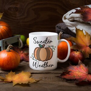 May include: White ceramic mug with a black and brown pumpkin design and the text "Sweater Weather". The mug is surrounded by fall foliage, including maple leaves and pumpkins.