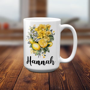 May include: White ceramic mug featuring a floral design with yellow lemons and flowers. The mug has the name "Hannah" in black script. The handle is on the right side of the mug.