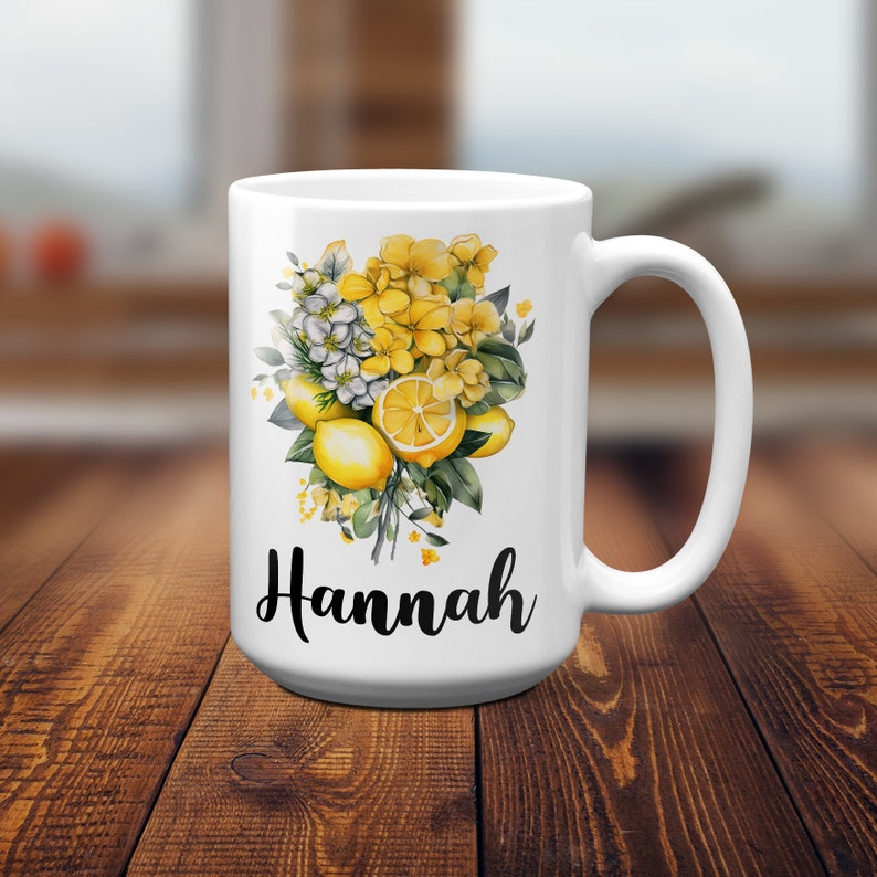 May include: White ceramic mug featuring a floral design with yellow lemons and flowers. The mug has the name "Hannah" in black script. The handle is on the right side of the mug.