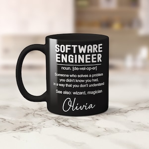 K&ouml;nnte beinhalten: Schwarze Kaffeetasse mit einem wei&szlig;en Design, auf dem "SOFTWARE ENGINEER" steht und der Begriff definiert wird als "Jemand, der ein Problem l&ouml;st, von dem man nicht wusste, dass man es hat, auf eine Weise, die man nicht versteht. Siehe auch: Zauberer, Magier." Die Tasse ist mit dem Namen "Olivia" personalisiert.