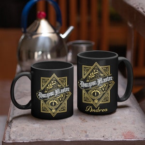 May include: Two black ceramic mugs with a gold design. The design features a stylized eye, a dice, and the text "Dungeon Master". One mug has the name "Andrea" printed below the design.