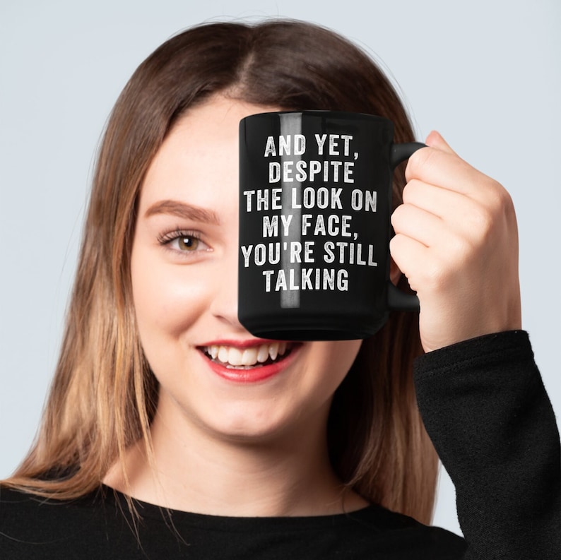 Pu&ograve; includere: Una tazza in ceramica nera con la scritta bianca: "AND YET, DESPITE THE LOOK ON MY FACE, YOU'RE STILL TALKING". La tazza &egrave; tenuta davanti al volto di una persona. La persona sta sorridendo.