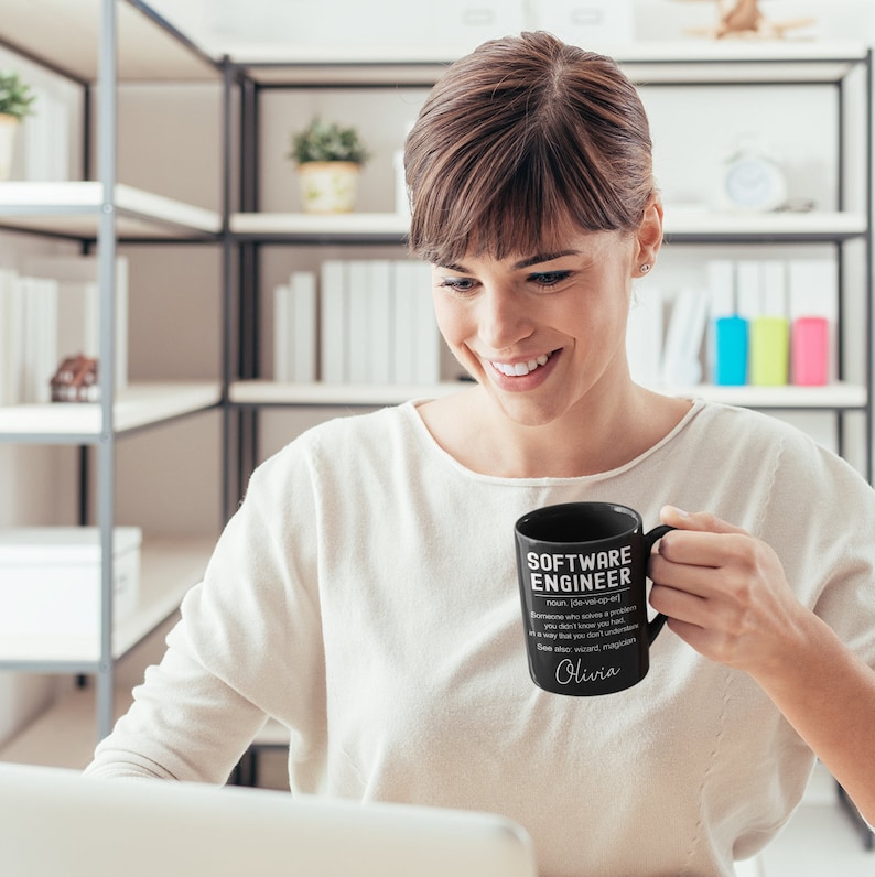 K&ouml;nnte beinhalten: Schwarze Kaffeetasse mit wei&szlig;em Text, der "Software Engineer" lautet und eine Definition des Begriffs enth&auml;lt. Die Tasse tr&auml;gt au&szlig;erdem den Namen "Olivia", der darauf gedruckt ist.