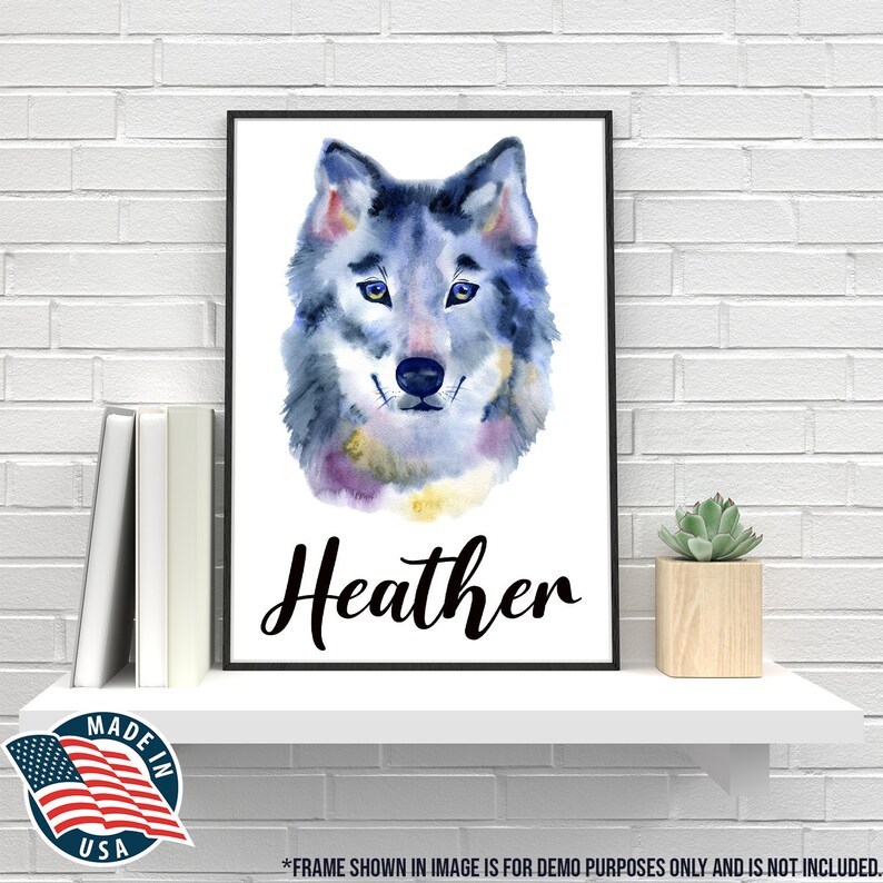 May include: A framed watercolor print of a wolf's face, predominantly blue and gray, with yellow eyes. The name "Heather" is written in black script below the image. The print is displayed on a white shelf with books and a small succulent.