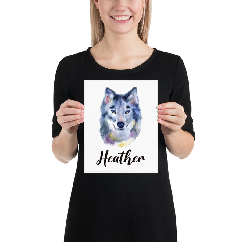May include: A watercolor print featuring a wolf's head with blue, gray, and yellow hues. The name "Heather" is written in cursive below the wolf illustration. The print is held by a person.