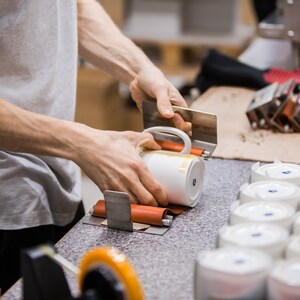 May include: A person is using a tool to apply a label to a white ceramic mug. The mug is being held in place by a metal clamp. There are several other mugs in the background.