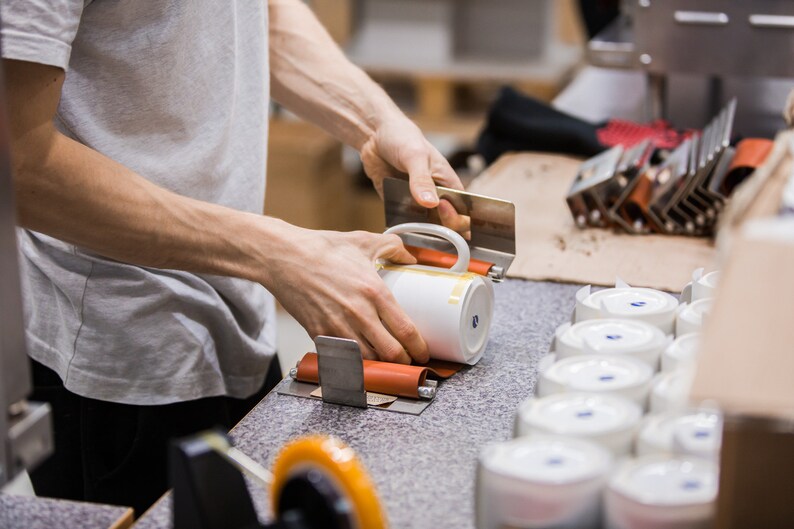 May include: A person is using a tool to apply a label to a white ceramic mug. The mug is being held in place by a metal clamp. There are several other mugs in the background.