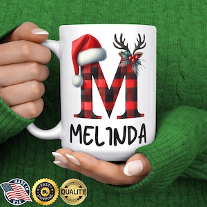 May include: White ceramic mug featuring a red and black buffalo plaid letter "M" with a Santa hat and deer antlers. The name "Melinda" is printed below the letter in black.