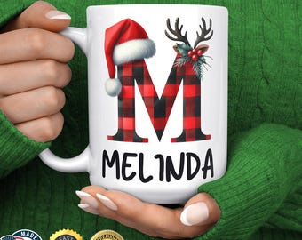 Könnte beinhalten: Weiße Keramiktasse mit einem rot-schwarz karierten Buchstaben "M" mit Weihnachtsmütze und Hirschgeweih. Der Name "Melinda" ist darunter in Schwarz gedruckt.
