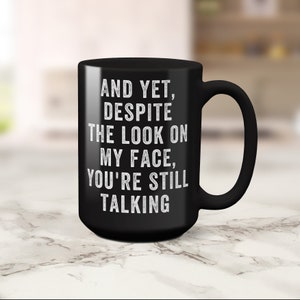 Pu&ograve; includere: Tazza da caff&egrave; in ceramica nera con manico curvo. La tazza presenta un testo bianco che recita: "AND YET, DESPITE THE LOOK ON MY FACE, YOU'RE STILL TALKING."