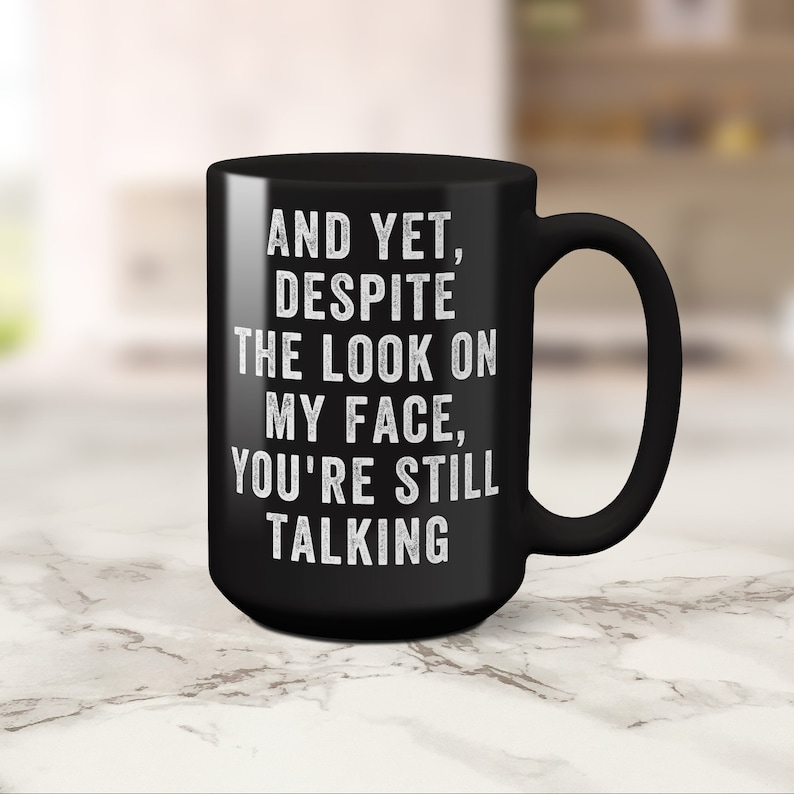 Pu&ograve; includere: Tazza da caff&egrave; in ceramica nera con manico curvo. La tazza presenta un testo bianco che recita: "AND YET, DESPITE THE LOOK ON MY FACE, YOU'RE STILL TALKING."