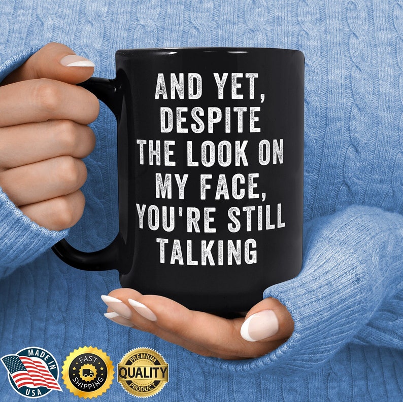 Pu&ograve; includere: Tazza in ceramica nera con la scritta bianca "AND YET, DESPITE THE LOOK ON MY FACE, YOU'RE STILL TALKING". La tazza &egrave; tenuta da una persona che indossa un maglione blu. La tazza ha un manico nero.