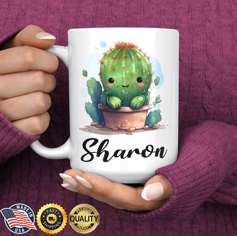 May include: White ceramic mug with a cartoon cactus illustration and the name "Sharon" printed on the front. The cactus is green with a red flower on top and is sitting in a brown pot.