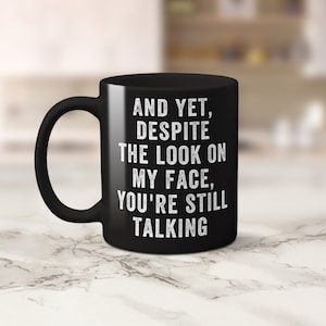 Pu&ograve; includere: Una tazza da caff&egrave; in ceramica nera con un manico curvo. La tazza presenta un testo bianco che recita: "E ANCORA, NONOSTANTE L'ASPETTO SULLA MIA FACCIA, STAI ANCORA PARLANDO."