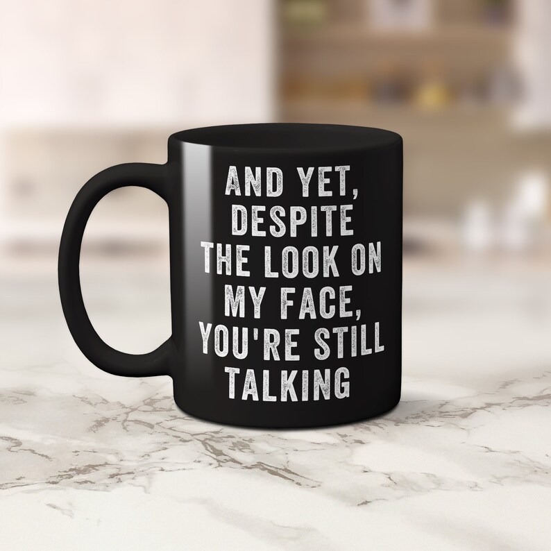 Pu&ograve; includere: Una tazza da caff&egrave; in ceramica nera con un manico curvo. La tazza presenta un testo bianco che recita: "E ANCORA, NONOSTANTE L'ASPETTO SULLA MIA FACCIA, STAI ANCORA PARLANDO."