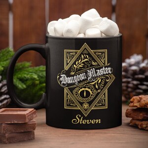 May include: Black ceramic mug with a gold design featuring a dragon eye and the text "Dungeon Master" with the name "Steven" below. The mug is filled with marshmallows.