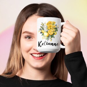 May include: White ceramic mug featuring a floral design with yellow hydrangeas, lemons, and green leaves. The name "Kelianne" is printed in black script. The mug has a large handle and is held up by a person.