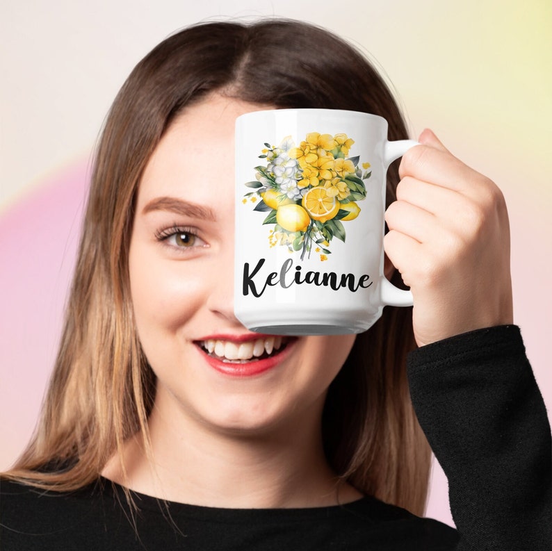 May include: White ceramic mug featuring a floral design with yellow hydrangeas, lemons, and green leaves. The name "Kelianne" is printed in black script. The mug has a large handle and is held up by a person.