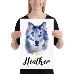 May include: A watercolor print featuring a wolf's head with blue, gray, and purple hues. The wolf has yellow eyes and a black nose. The name "Heather" is written in cursive below the image. The print is held by a person.