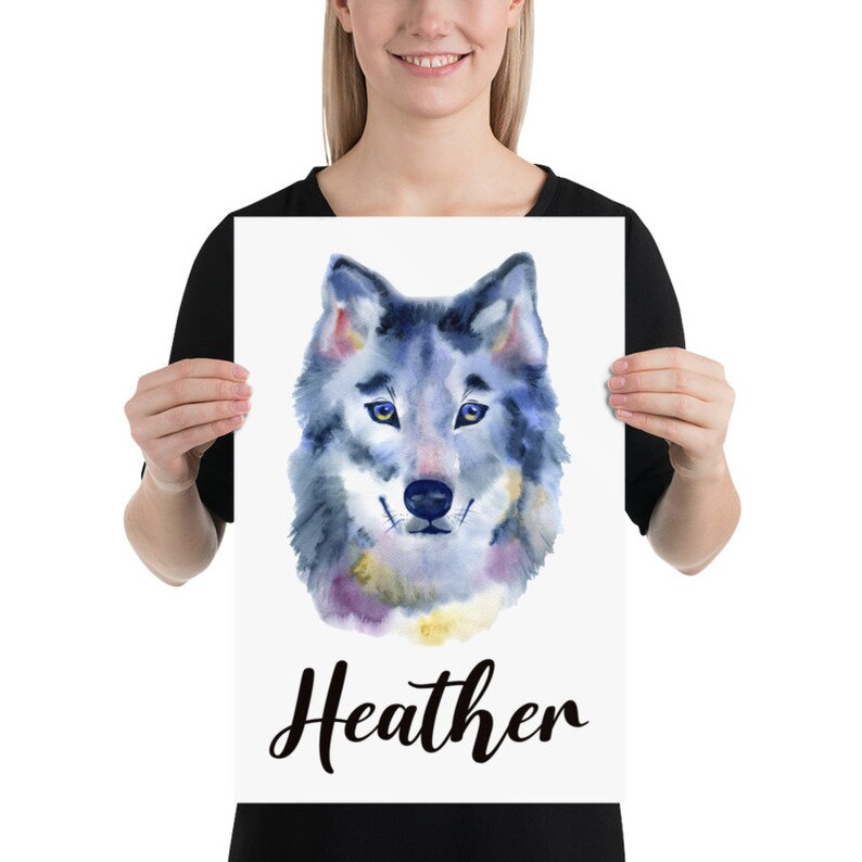 May include: A watercolor print featuring a wolf's head with blue, gray, and purple hues. The wolf has yellow eyes and a black nose. The name "Heather" is written in cursive below the image. The print is held by a person.