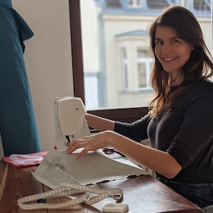 May include: A woman smiles while using a sewing machine at a wooden desk near a window. A roll of teal fabric leans against the wall. A measuring tape and a small white bar of soap are on the desk. The woman is wearing a dark gray long-sleeve shirt.