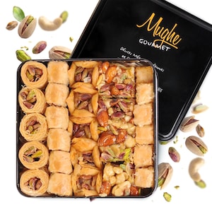 May include: A square black tin box labeled Mughe Gourmet filled with assorted baklava pastries, including shredded dough nests with pistachios, layered phyllo squares, and nut-topped pieces, surrounded by scattered pistachios and cashews.