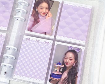 May include: A white binder with clear plastic sleeves containing two rectangular photo cards. One card shows a person with long brown hair, and the other shows a person with a purple hair bow holding a small blue and pink carousel toy.