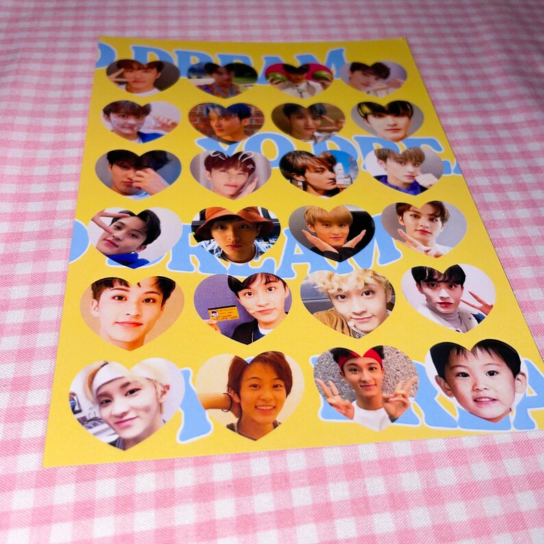 Op de afbeelding: Een geel vel papier met hartvormige stickers met foto's van een boyband. De stickers zijn in een raster gerangschikt en elk hart heeft een blauwe omtrek. De tekst "DREAM YOU ARE DREAM" is in blauwe letters gedrukt boven en onder op het vel.