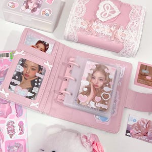 Peut inclure: Un album photo rose avec un mécanisme à anneaux, ouvert pour afficher des photos. L'album est entouré d'autocollants, d'une petite boîte transparente avec une photo et d'un portefeuille rose avec une garniture en dentelle. L'album a une fermeture en forme de cœur.