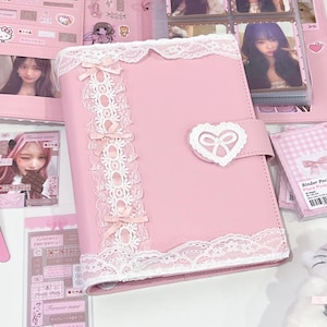 May include: A pink binder with a heart-shaped clasp and white lace trim. The binder features small pink bows and is surrounded by other pink accessories, including photo sleeves and stickers.