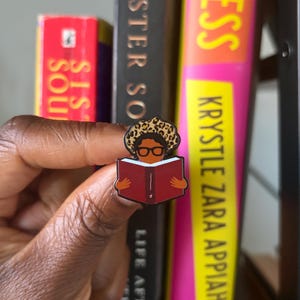 May include: A metal enamel pin featuring a stylized figure with dark skin, wearing black-rimmed glasses and a leopard-print head covering, holding an open red book. The figure is depicted from the chest up with a simple, graphic design style.
