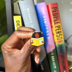 May include: A metal enamel pin featuring a person with dark skin and dark hair reading an open yellow book. The book has a small red square detail on the left page and a light green rectangle on the right page. The pin has a black outline.