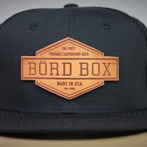 Puede incluir: Gorra de béisbol negra y marrón con un parche de cuero que dice "The First Portable Skateboard Rack" y "BÖRD BOX™ Made in USA Pat. Pend."