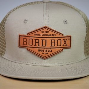 Puede incluir: Gorra trucker beige con parte trasera de malla y visera plana. Cuenta con un parche de cuero marrón con las palabras "BÖRD BOX" y "MADE IN USA". El parche también dice "THE FIRST PORTABLE SKATEBOARD RACK".