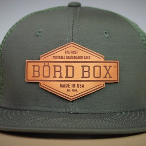 Könnte beinhalten: Eine grüne und schwarze Mesh-Baseballkappe mit einem braunen Leder-Patch, auf dem "The First Portable Skateboard Rack" und "BÖRD BOX™" mit dem Text "Made in USA Pat. Pend." steht.