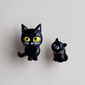 May include: Two black cat figurines. The larger cat has yellow eyes and a white moustache. The smaller cat has white eyes and a gold collar.